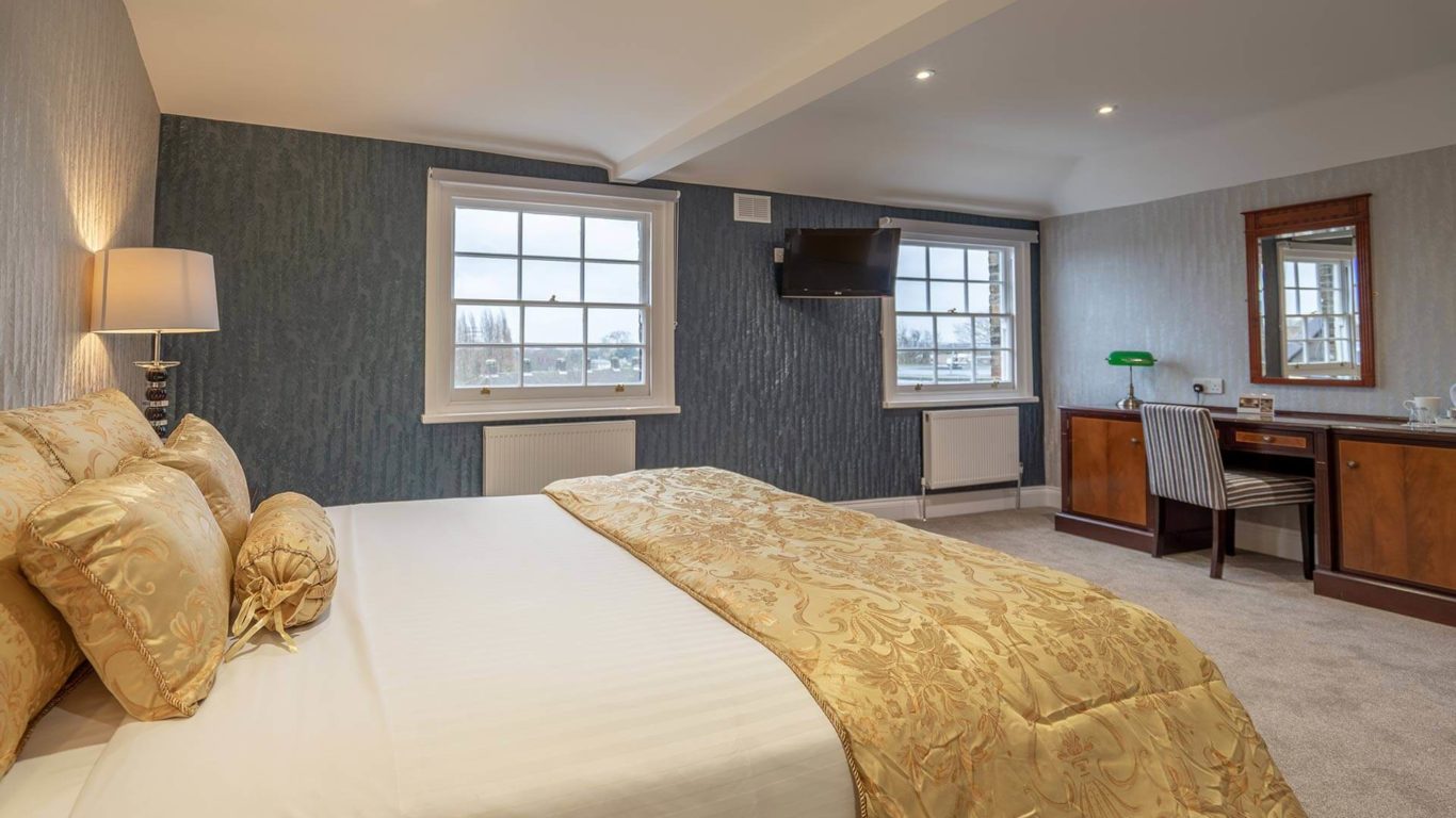 Elegant hotel room with plush gold bedding, desk, flat-screen TV, and large windows offering scenic views. Welcoming and cosy.