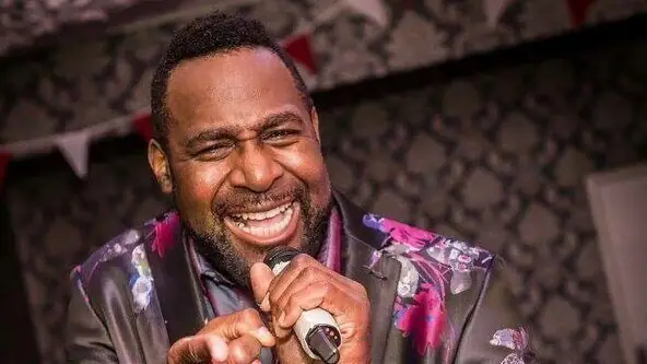 A smiling performer sings into a microphone at The Clarendon, exuding joy in a lively, patterned setting.