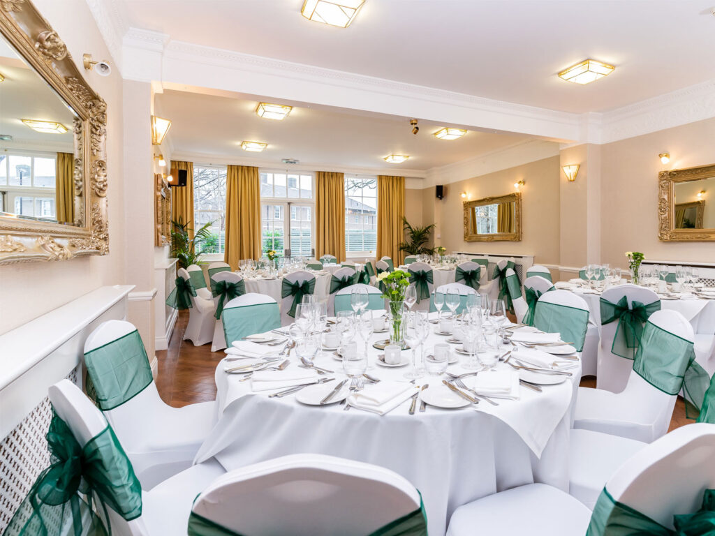 Charming Event Space at The Clarendon Elegant dining room with round tables, white tablecloths, green sashes on chairs, mirrors, and large windows at The Clarendon Hotel.