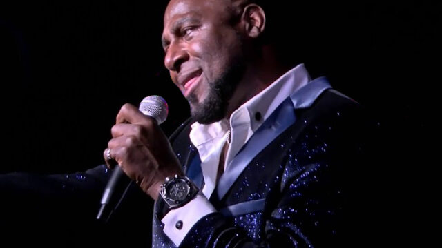 Man joyfully singing on stage with microphone, wearing a sparkling navy jacket, evoking lively entertainment atmosphere.
