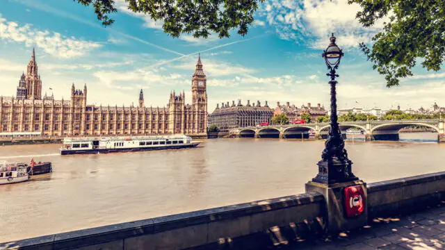 River Thames view with lush trees, iconic Houses of Parliament, and serene riverboats; perfect for a relaxing London stay.